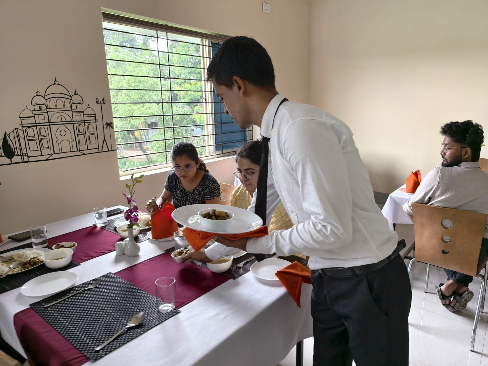 A student presenting a dish on a service platter to guests seated at a laid table in the SHM training restaurant