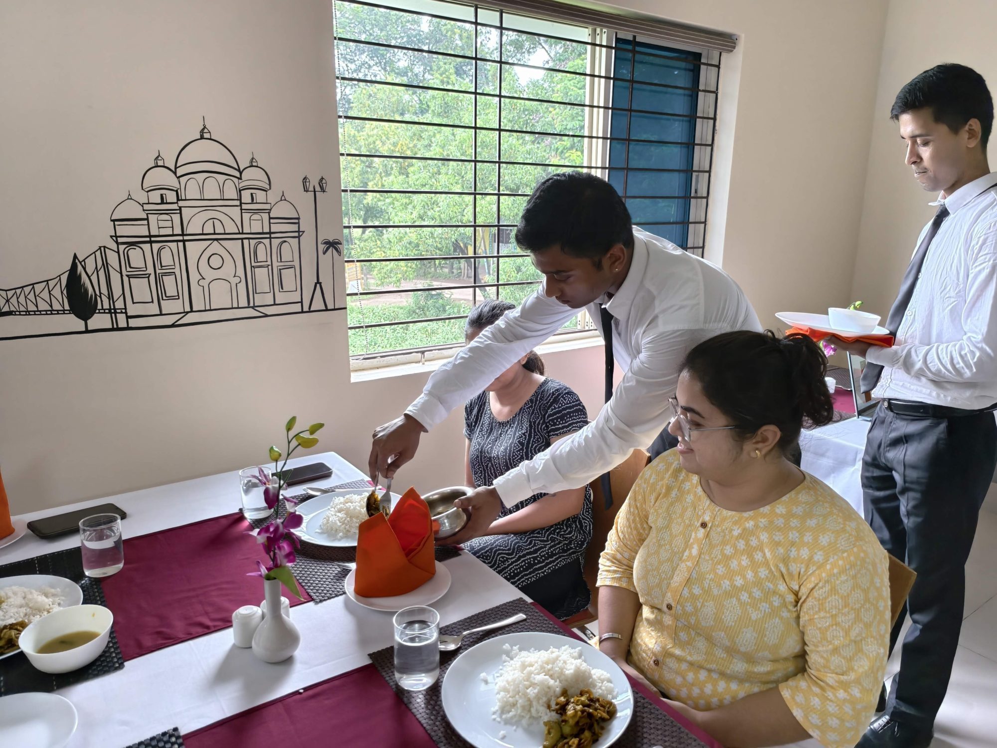 A student practising silver service, serving a guest from a silver bowl at a laid table in the SHM training restaurant