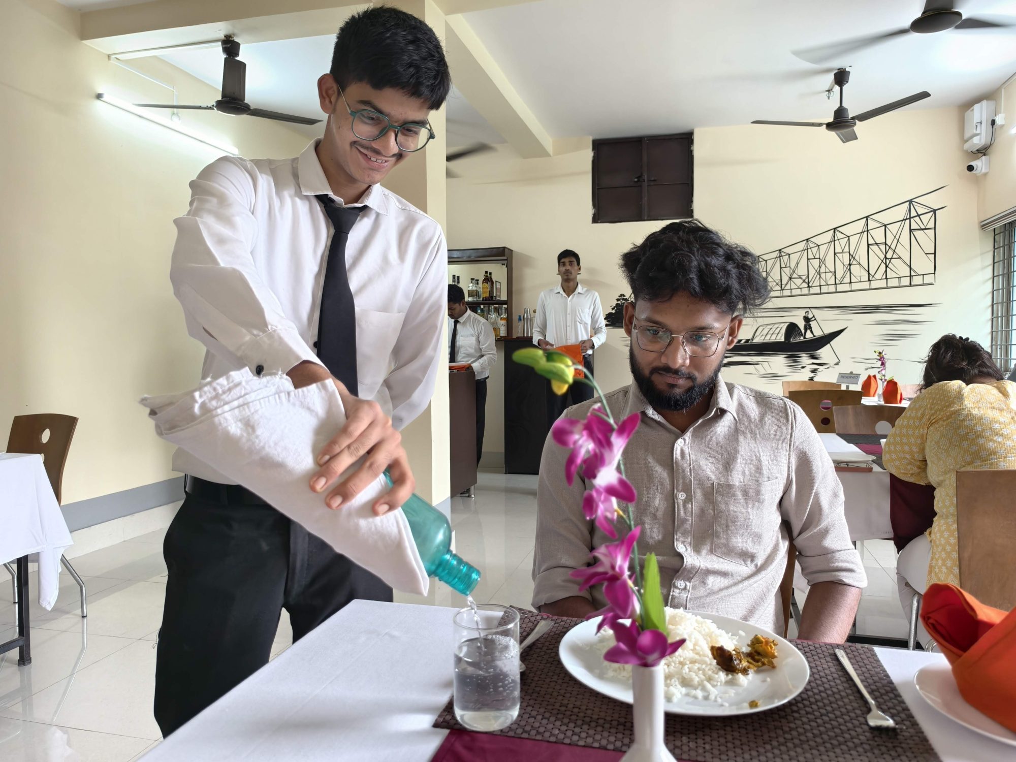 A student pouring water from a napkin-wrapped bottle, practising water service at SHM Durgapur